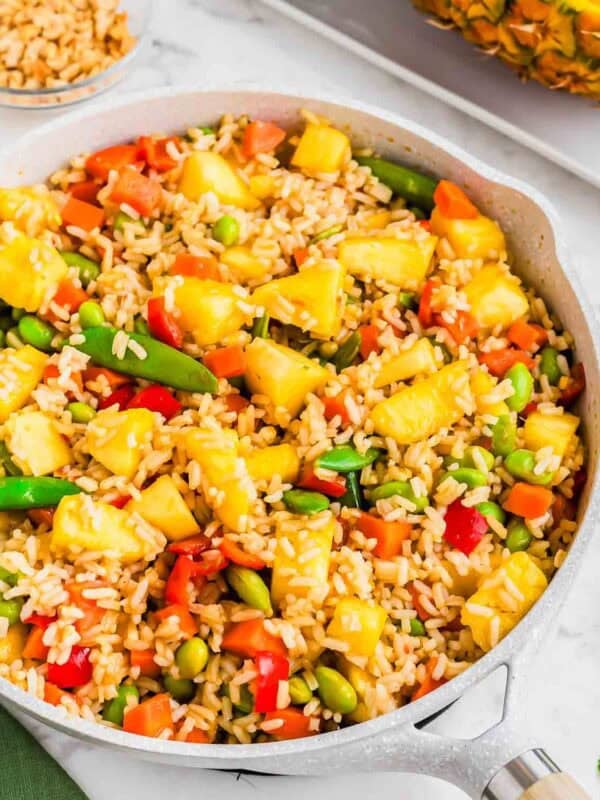 A skillet filled with a colorful fried rice dish featuring pineapple chunks, red bell peppers, edamame, snap peas, and carrots. A bowl of chopped peanuts and a half pineapple are in the background on a white surface.