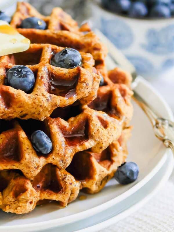 A stack of sweet potato waffles topped with fresh blueberries and a pat of butter on a white plate. A vintage silver fork rests beside. In the background, there's a bowl of blueberries and a dish of syrup.