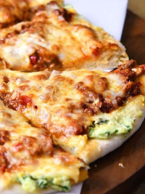 A close-up of a pizza with melted cheese and toppings on a wooden board. The pizza is sliced into servings and partially visible. In the background, there's a jar of chili flakes and a jar of grated cheese.