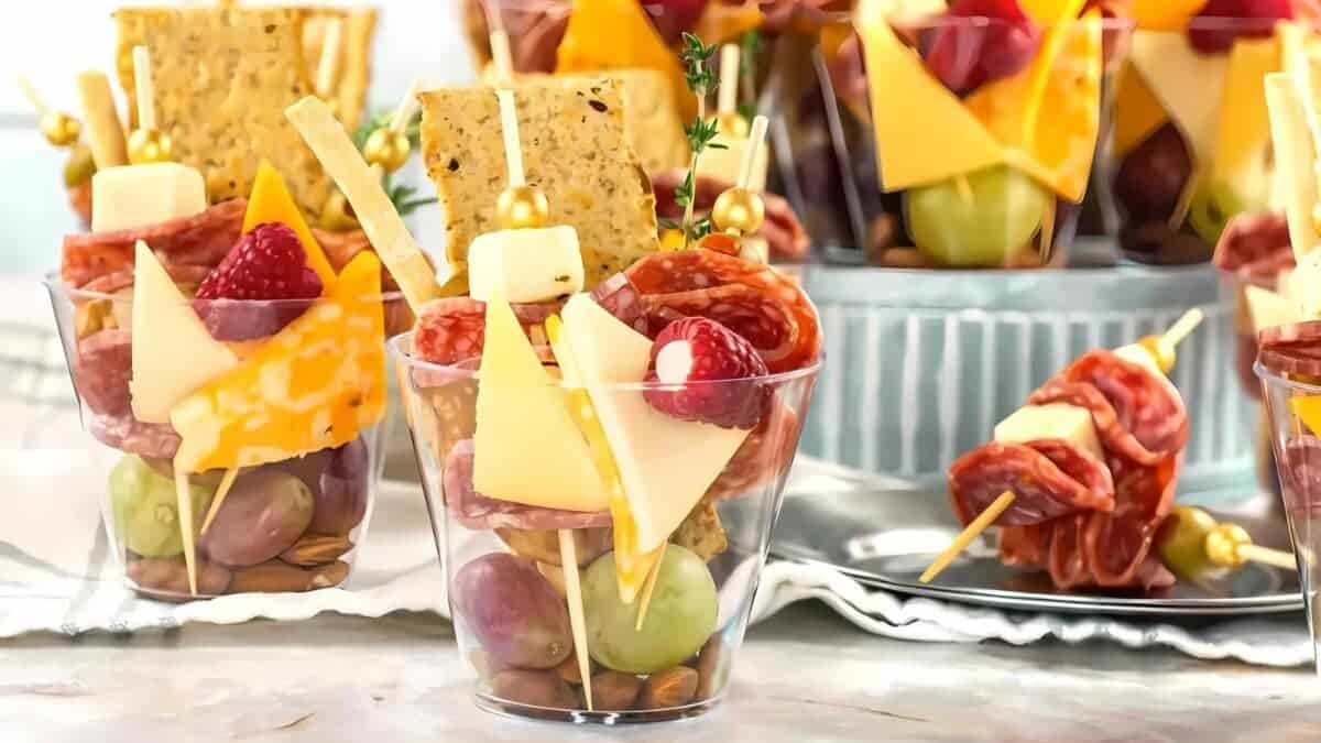 Clear plastic cups filled with assorted snacks including grapes, cheese cubes, meat slices, skewered cheese and fruit, crackers, and nuts, arranged neatly for a party or gathering.