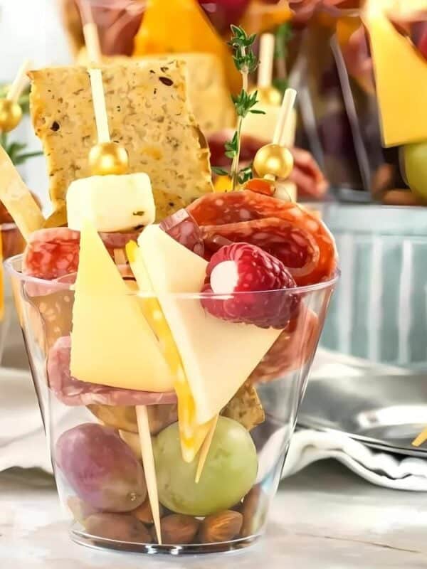 Clear plastic cups filled with assorted snacks including grapes, cheese cubes, meat slices, skewered cheese and fruit, crackers, and nuts, arranged neatly for a party or gathering.