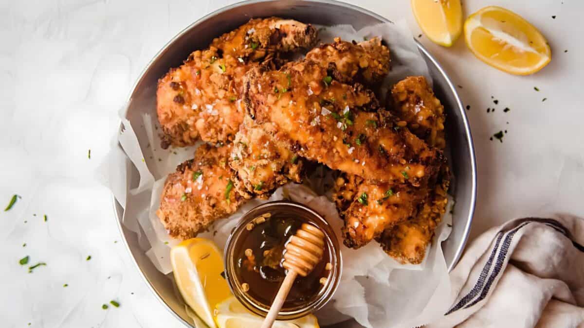 A bowl of crispy fried chicken tenders garnished with chopped herbs, served with lemon wedges and a small dish of honey, with a honey dipper resting inside. A striped napkin is partially visible on the side.