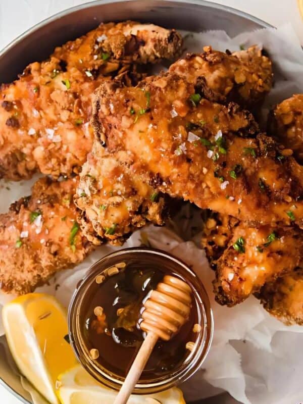 A bowl of crispy fried chicken tenders garnished with chopped herbs, served with lemon wedges and a small dish of honey, with a honey dipper resting inside. A striped napkin is partially visible on the side.