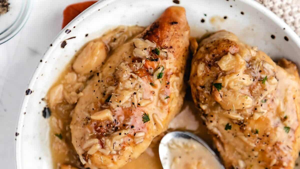 A white plate with two pieces of cooked chicken breast, covered in a creamy garlic sauce and garnished with herbs. A spoon rests in the sauce beside the chicken.