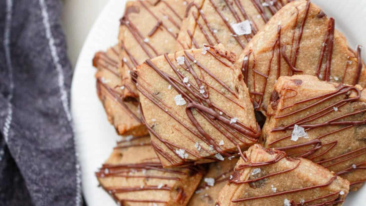 A plate of golden-brown, triangular cookies drizzled with chocolate and sprinkled with flaky sea salt. The cookies are arranged in an overlapping pattern. A dark cloth napkin is partially visible on the left side.