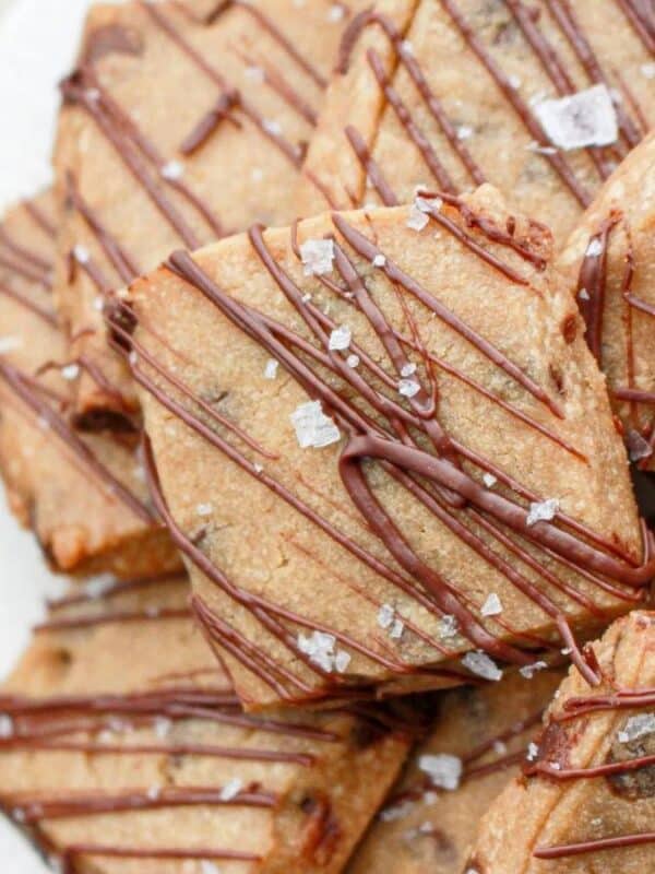 A plate of golden-brown, triangular cookies drizzled with chocolate and sprinkled with flaky sea salt. The cookies are arranged in an overlapping pattern. A dark cloth napkin is partially visible on the left side.
