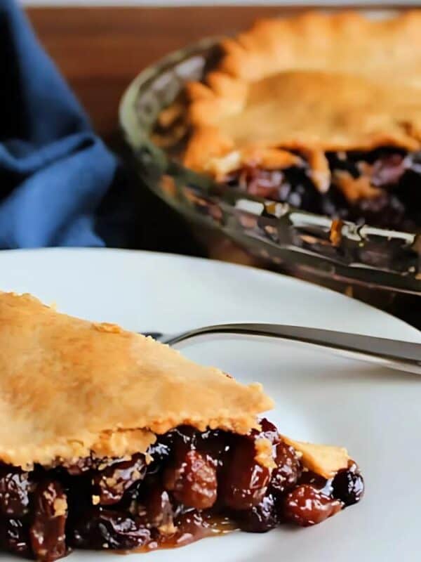 A slice of raisin pie on a white plate with a fork, sitting in front of the remaining pie in a glass dish. A blue cloth napkin is in the background on a wooden table.