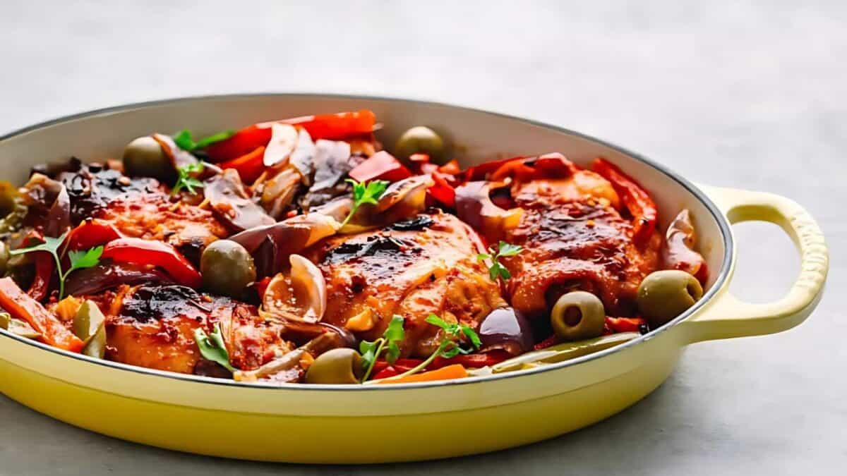 A yellow dish filled with baked chicken topped with a tomato-based sauce, surrounded by green olives, sliced red peppers, mushrooms, and garnished with fresh parsley.