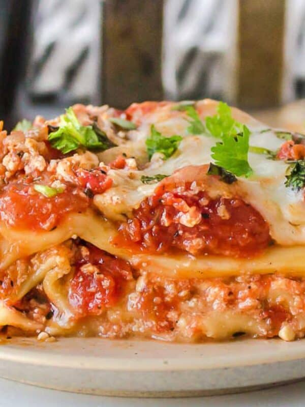 A close-up of a slice of lasagna on a beige plate. The dish is layered with pasta, tomato sauce, ground meat, melted cheese, and topped with parsley. A blurred background features another plate of lasagna.