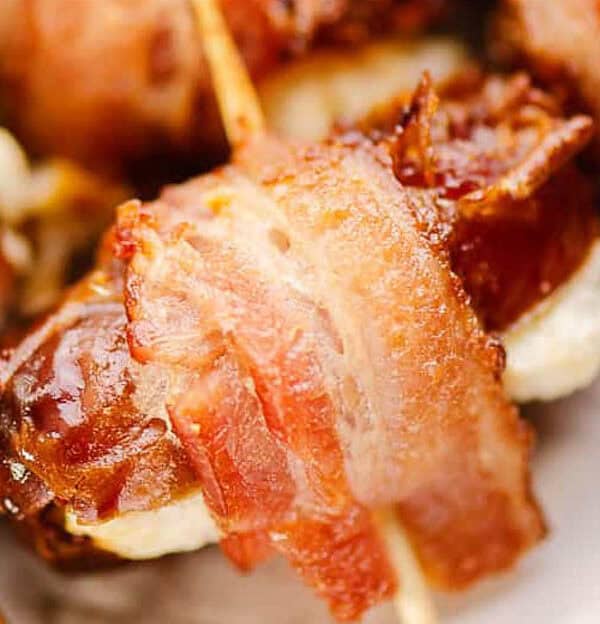 Close-up of bacon-wrapped dates with cream cheese filling, secured with toothpicks. They are served on a light-colored plate, highlighting the crispy texture of the bacon and creamy filling.