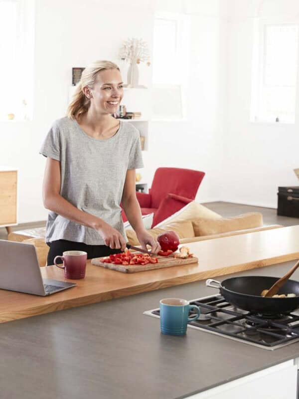 A woman and a man are in a modern kitchen. The woman is slicing tomatoes on a wooden board near a laptop. The man is cooking on a stove, stirring a pan. They are smiling at each other. The room has a TV, shelves, and bright decor.