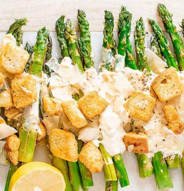 Grilled asparagus spears topped with Caesar dressing, parmesan cheese, and croutons, garnished with lemon halves. A bowl of extra croutons is on the side.