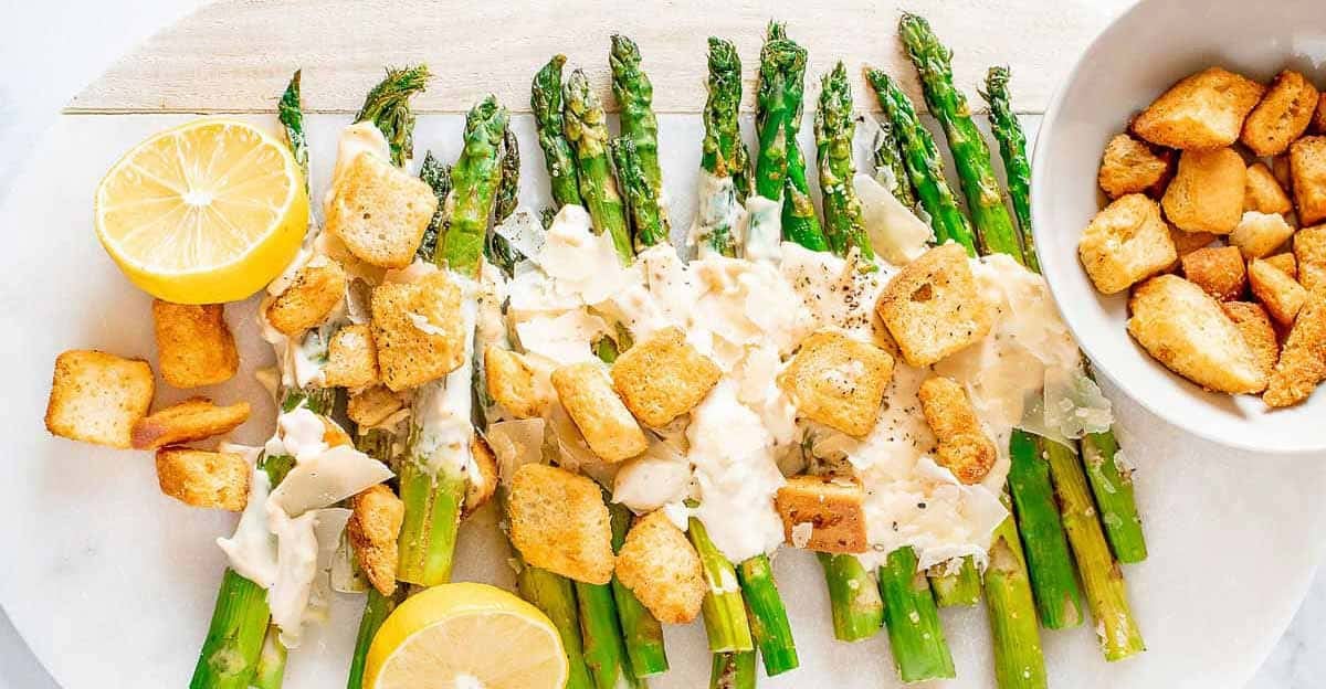 Grilled asparagus spears topped with Caesar dressing, parmesan cheese, and croutons, garnished with lemon halves. A bowl of extra croutons is on the side.