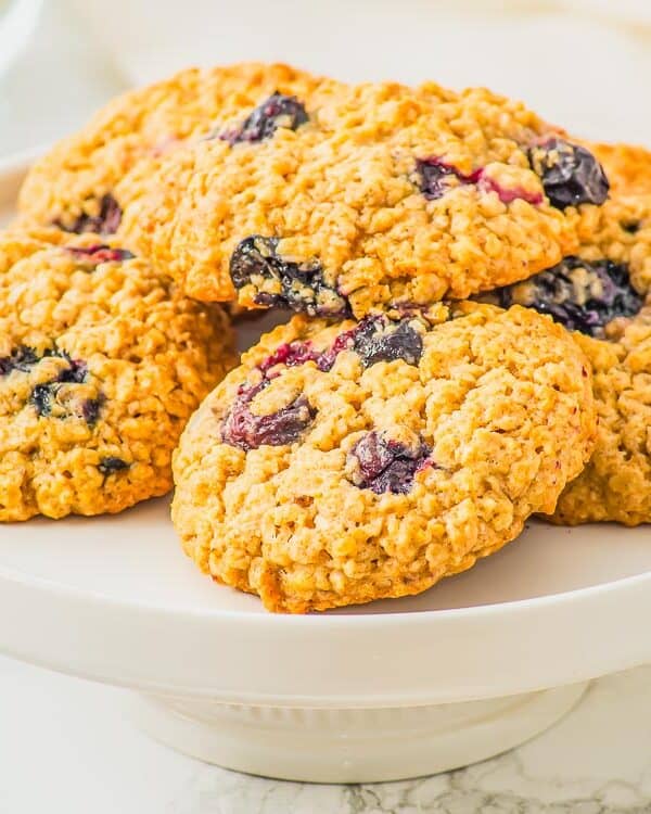 A white plate displays a stack of blueberry oatmeal cookies. In the background, there are fresh blueberries in a bowl, all set on a marble surface. The scene is brightly lit, highlighting the cookies' texture.