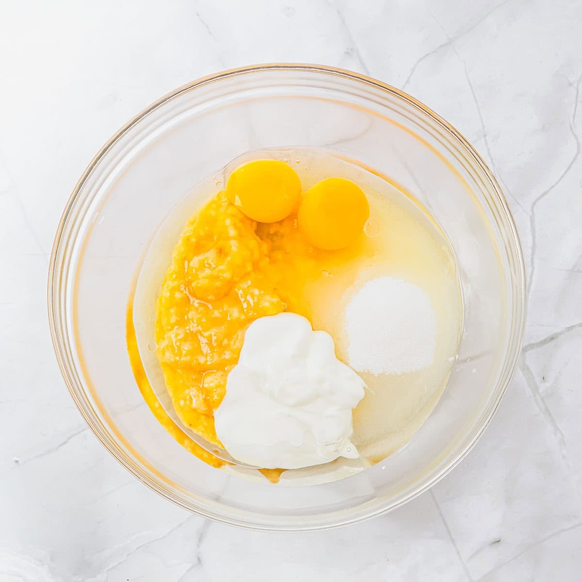 A glass bowl with mashed banana, two raw eggs, sugar, yogurt, and oil on a white marble surface, ready to be mixed for baking.
