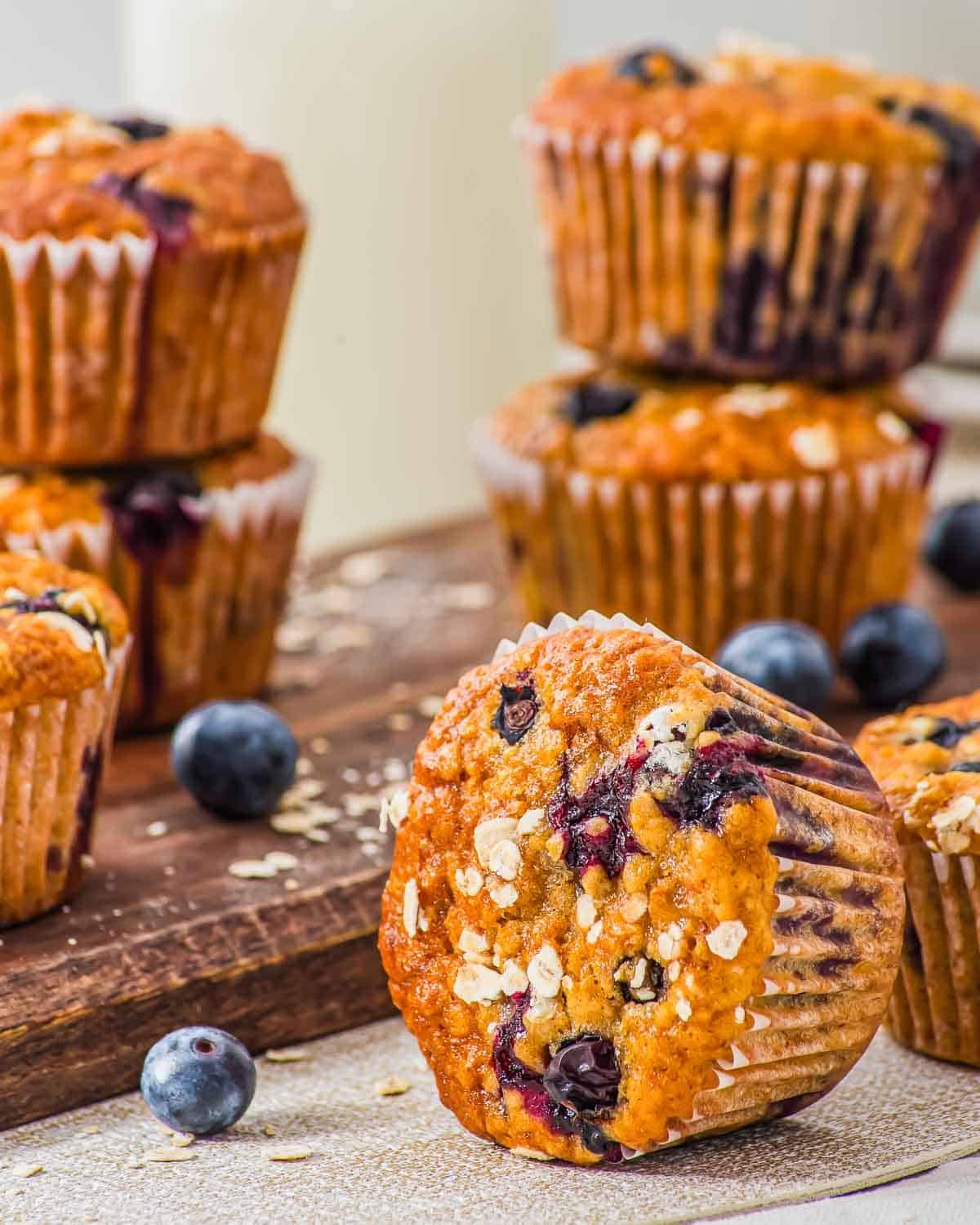 A close-up of oatmeal blueberry muffins topped with oats, arranged on a wooden board with fresh blueberries scattered around. One muffin is tilted forward, revealing its baked texture and blueberry filling.