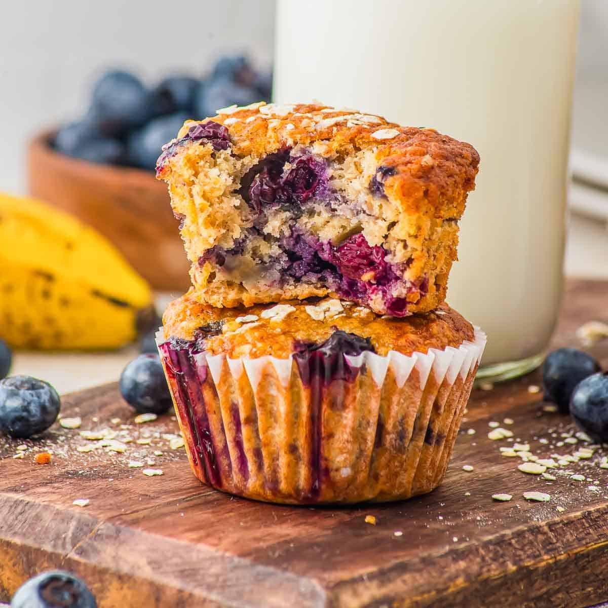 Two blueberry oat muffins sit on a wooden board, one stacked on top of the other with a bite taken out of the top muffin. Fresh blueberries, a banana, and a glass of milk are in the background.