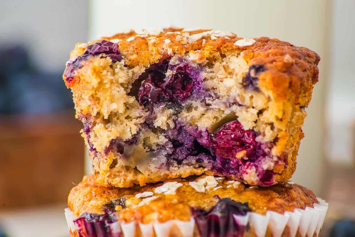Close-up of a blueberry banana oat muffin cut in half, showing a moist interior with visible juicy blueberries and oatmeal on top, placed on top of another whole muffin with a white paper liner.