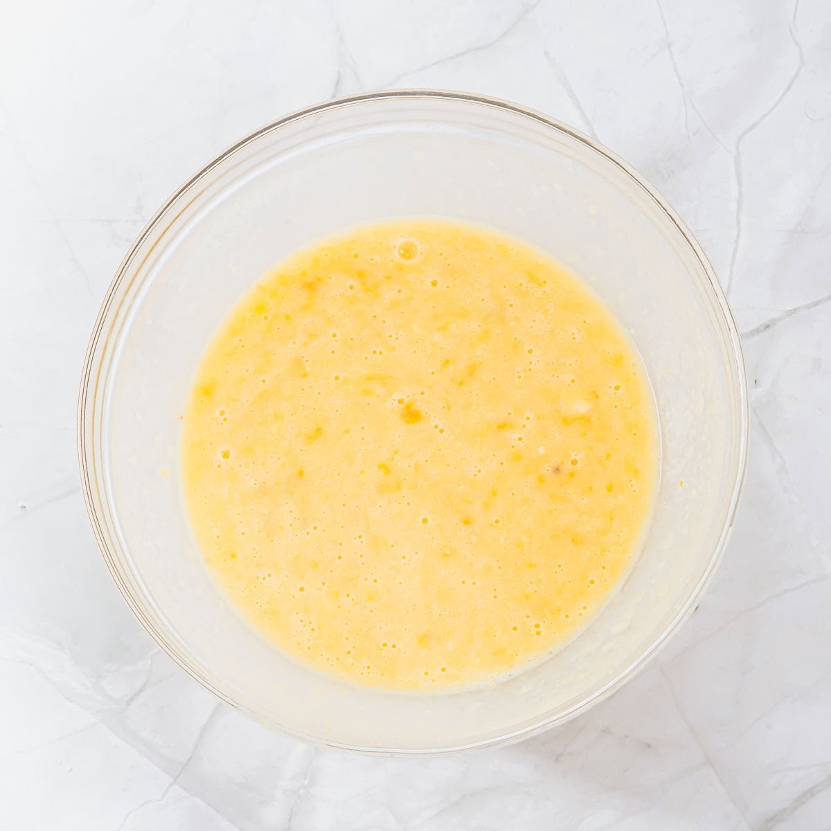 A clear glass bowl filled with a yellow, slightly lumpy batter sits on a white marble countertop.
