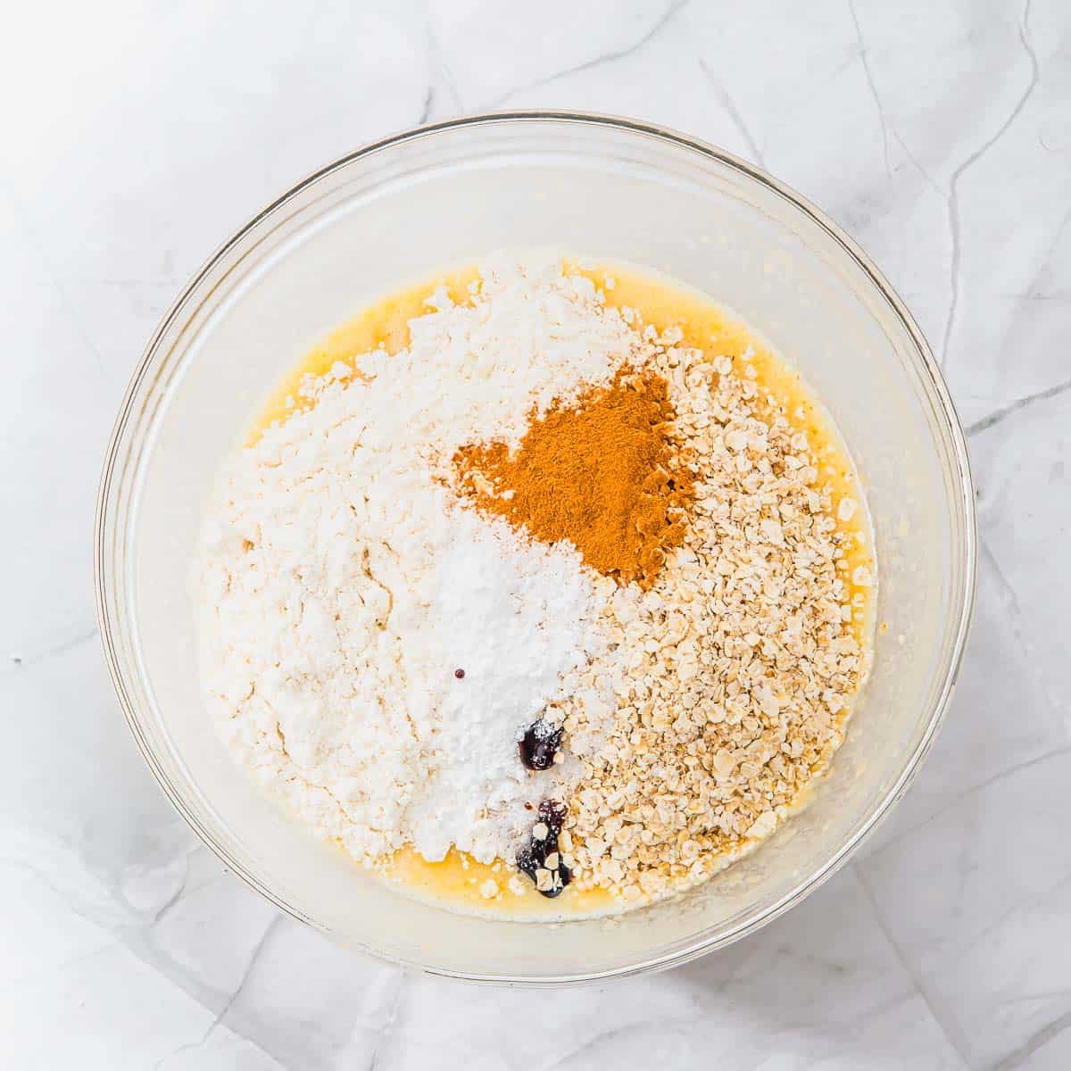 A glass bowl filled with flour, oats, sugar, cinnamon, and some liquid ingredients, placed on a white marble surface. The ingredients are not yet mixed together.