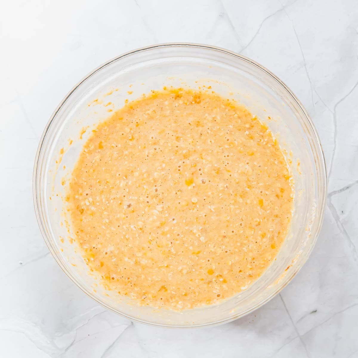 A clear glass bowl filled with a thick, yellowish muffin batter sits on a white marble surface.