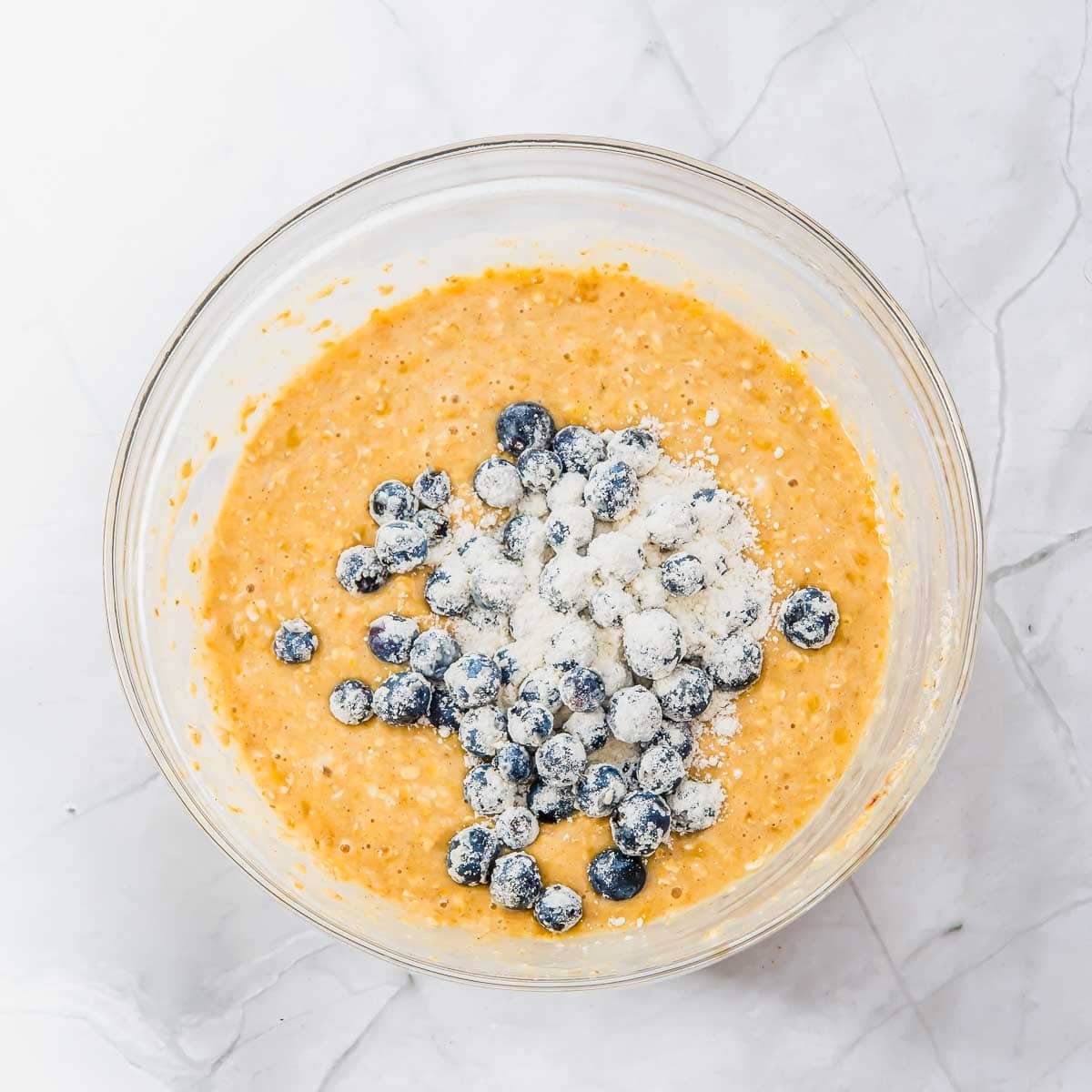 A glass bowl filled with yellowish muffin batter, topped with fresh blueberries dusted in flour, sits on a white marble surface.