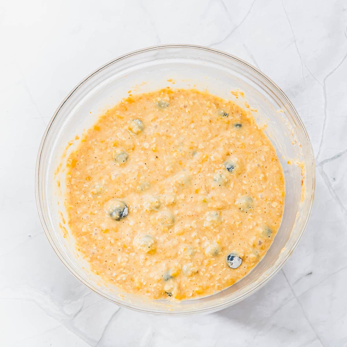 A clear glass bowl filled with a thick, orange-yellow muffin batter containing visible whole blueberries, sitting on a white marble surface.