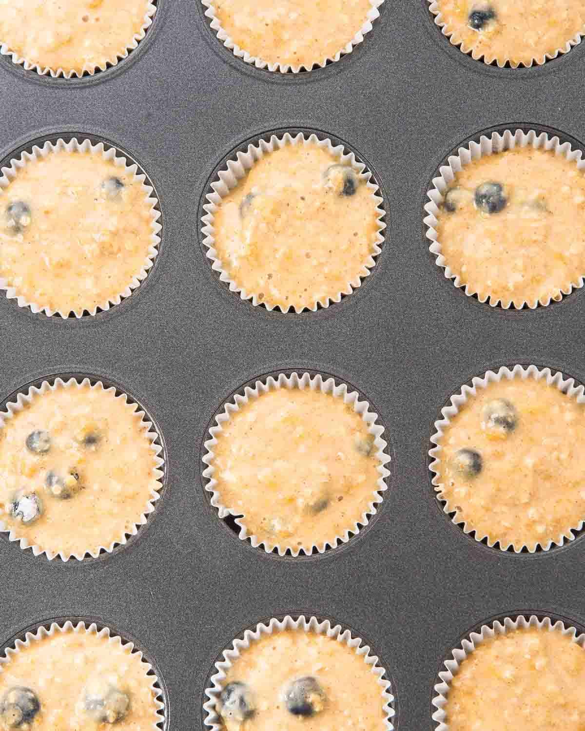 A muffin tin filled with paper liners, each holding unbaked blueberry muffin batter with visible blueberries. The batter is light tan in color and the pan is viewed from above.