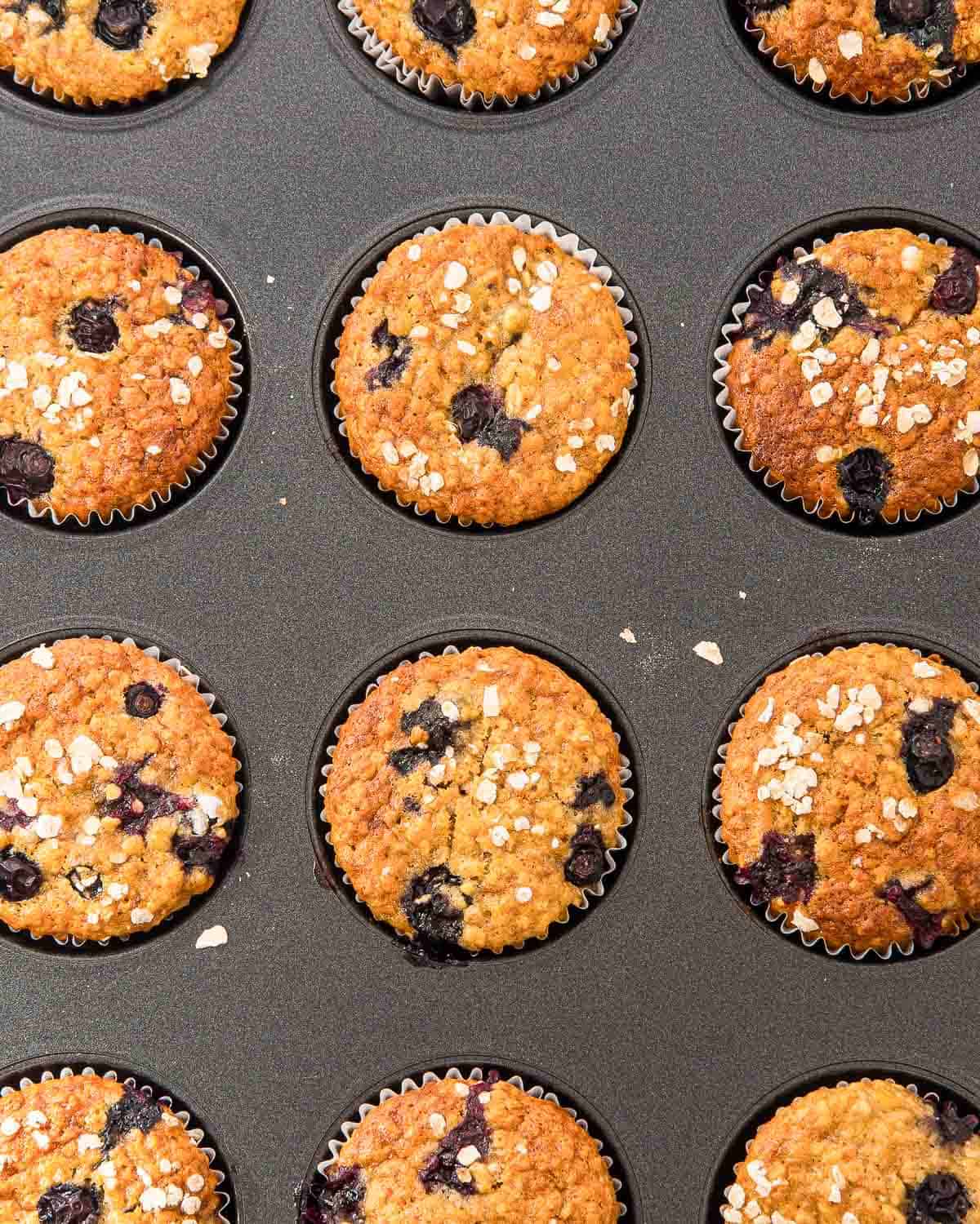A muffin tin filled with golden-brown blueberry muffins, each topped with oats and visible blueberry pieces. The muffins are baked in paper liners and arranged neatly in the pan.