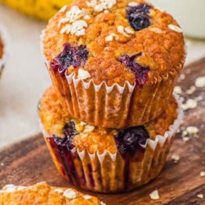 Two blueberry oatmeal muffins in paper liners are stacked on a wooden surface. The muffins are golden-brown with visible blueberries and oats on top. A banana and a third muffin are partially visible in the background.