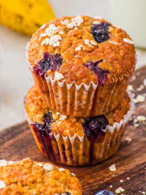 Two blueberry oatmeal muffins in paper liners are stacked on a wooden surface. The muffins are golden-brown with visible blueberries and oats on top. A banana and a third muffin are partially visible in the background.