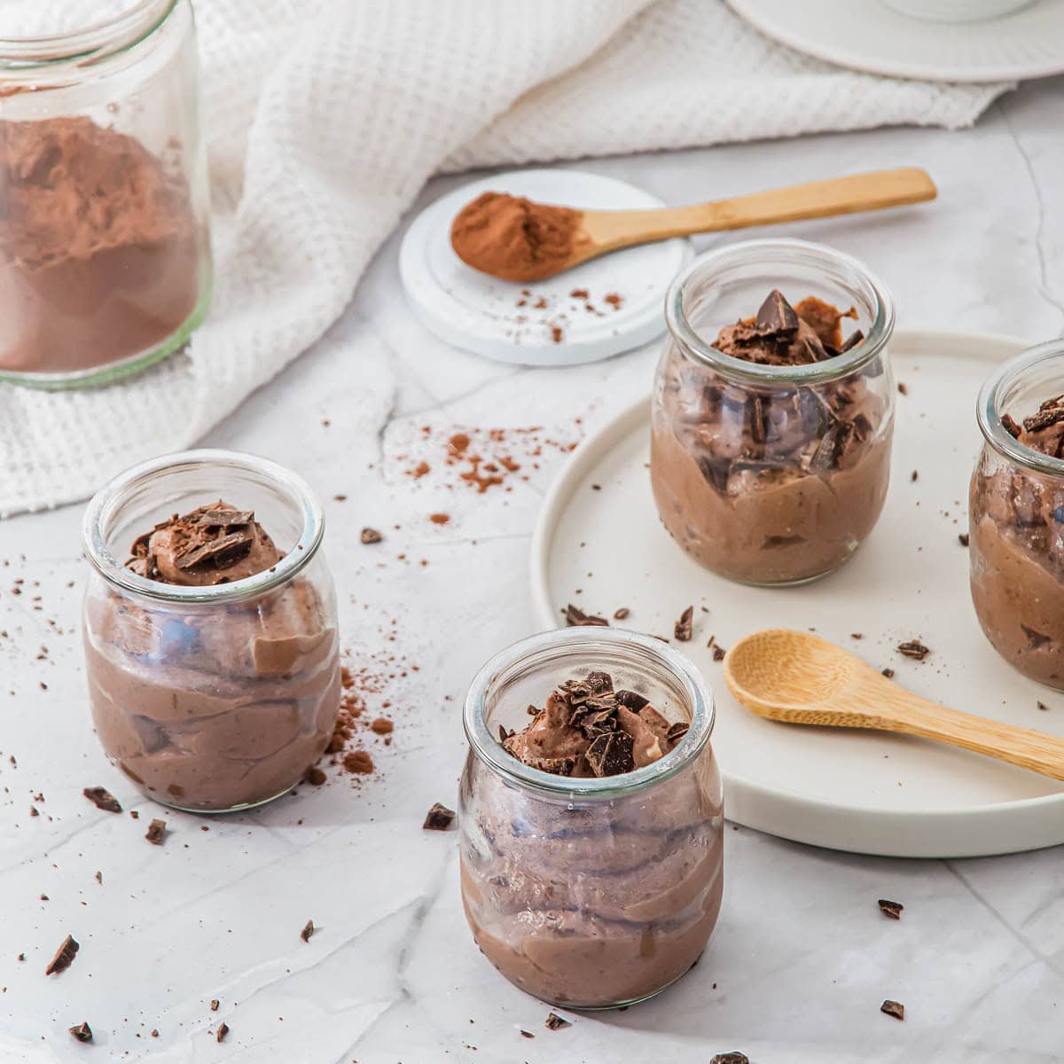 Four glass jars filled with chocolate mousse topped with chocolate shavings are placed on a marble surface. A wooden spoon rests on a white tray nearby, and cocoa powder is scattered around. A jar of cocoa is in the background.