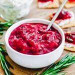 A white bowl filled with plum jam, placed on a wooden surface. A silver spoon rests in the bowl. Fresh rosemary sprigs lie nearby, and blurred crackers with cheese and cranberry sauce are in the background.