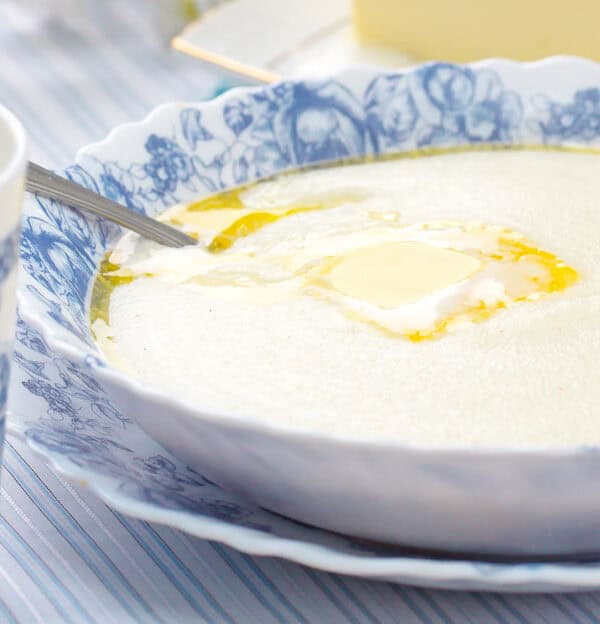 A bowl of semolina porridge topped with a pat of melting butter on a blue floral-patterned plate. Next to it is a cup of milk with a matching pattern, on a striped tablecloth.
