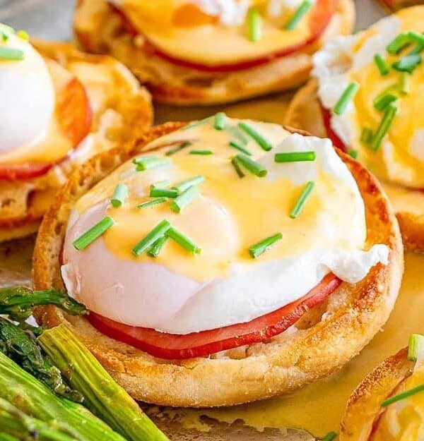 A close-up of eggs Benedict on English muffins with ham, poached eggs, and hollandaise sauce, garnished with chopped chives, served alongside roasted asparagus.