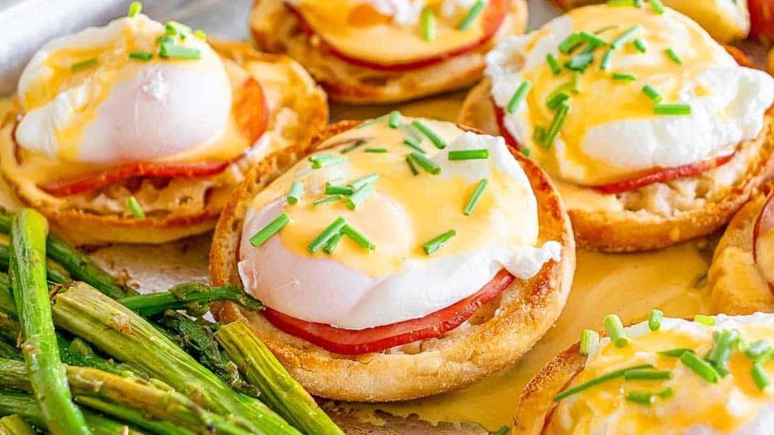 A close-up of eggs Benedict on English muffins with ham, poached eggs, and hollandaise sauce, garnished with chopped chives, served alongside roasted asparagus.