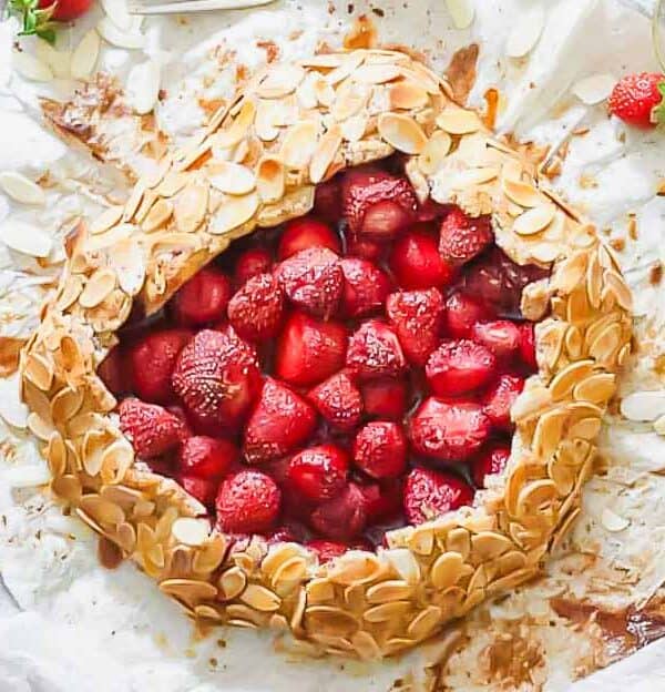 A rustic strawberry galette topped with sliced almonds is presented on a white cloth. Fresh strawberries are scattered around, along with a few almond slices. Two forks are placed nearby for serving.