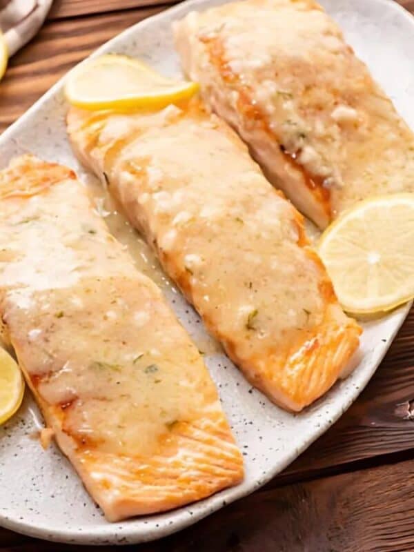 Three cooked salmon fillets with creamy sauce are served on a white oval plate, garnished with lemon slices. Fresh herbs and a brown-and-white cloth are nearby on a wooden table.