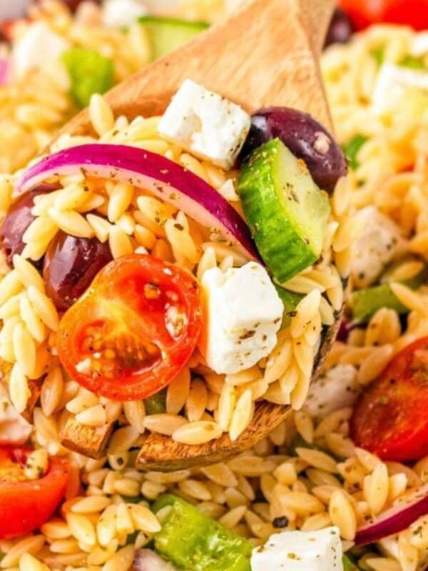 A wooden spoon scoops a colorful orzo pasta salad with cherry tomatoes, cucumber, red onion, kalamata olives, and feta cheese from a white bowl.