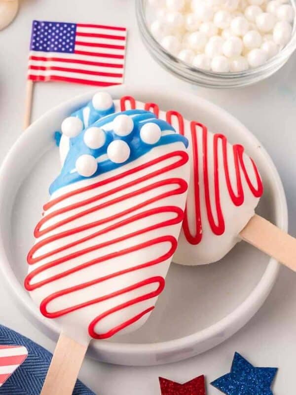 Patriotic ice cream pops decorated with red, white, and blue stripes and dots, mimicking the American flag, surrounded by small U.S. flags, blue stars, and a bowl of white candy pearls.