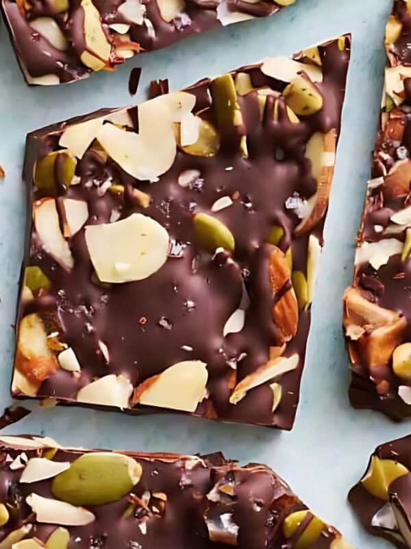 Pieces of dark chocolate bark topped with sliced almonds, pumpkin seeds, and other mixed nuts, arranged on a light blue surface.