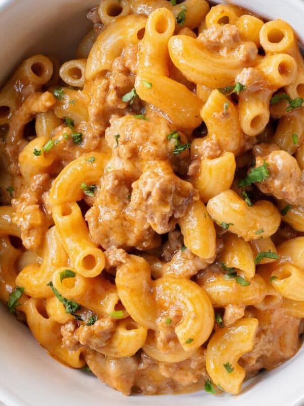 A white bowl filled with creamy macaroni and cheese mixed with ground meat and garnished with chopped herbs, placed on a light-colored surface next to a spoon and a napkin.