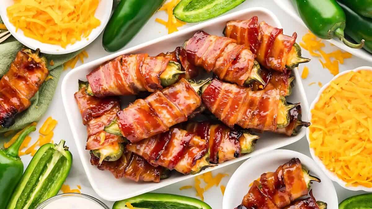 A platter of bacon-wrapped jalapeño poppers, garnished with melted cheese, surrounded by sliced jalapeños, shredded cheddar cheese, and dipping sauce on a white surface.