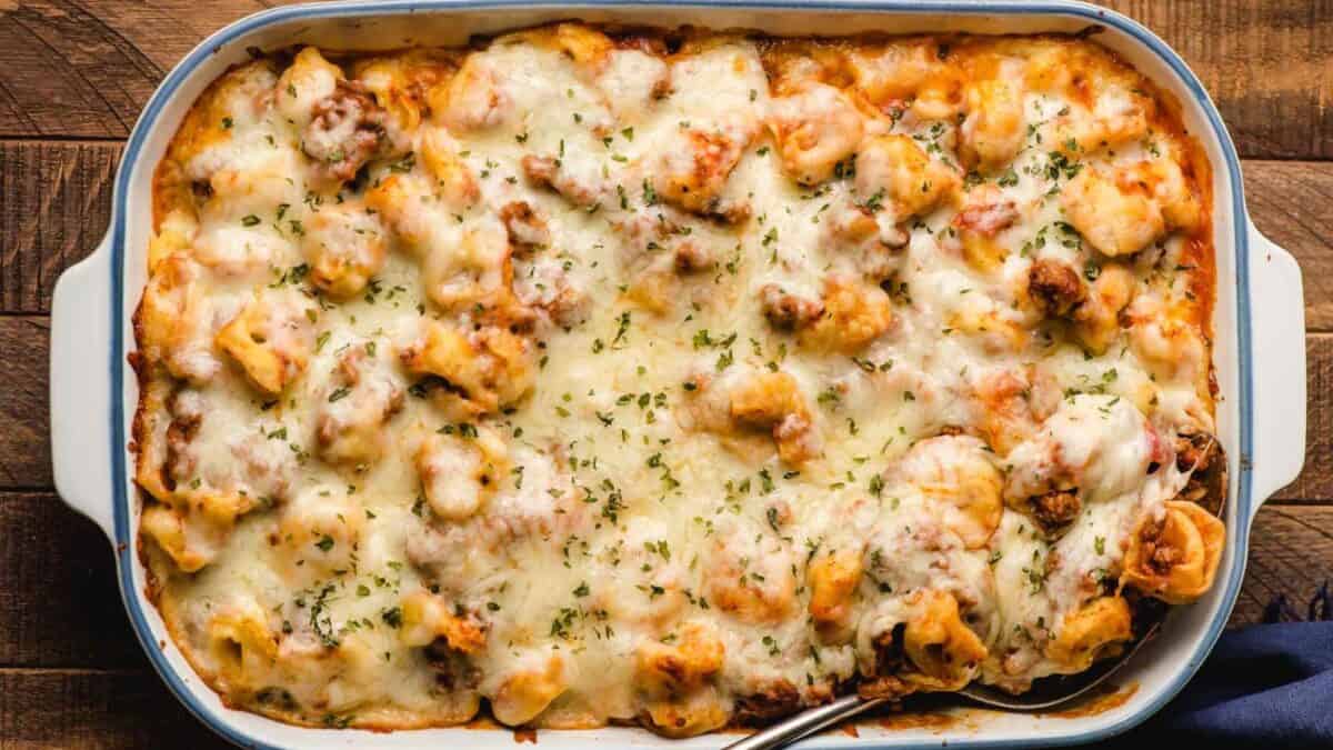A baked pasta casserole topped with melted, golden-brown cheese and sprinkled with herbs, served in a rectangular white dish on a wooden table.