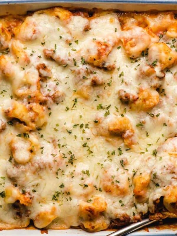 A baked pasta casserole topped with melted, golden-brown cheese and sprinkled with herbs, served in a rectangular white dish on a wooden table.