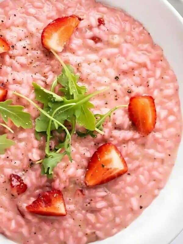 A white bowl filled with creamy pink risotto, garnished with fresh strawberry slices and arugula leaves, set on a light-colored table.