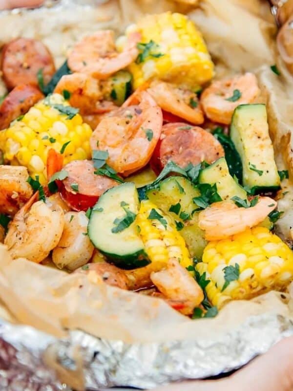 A close-up of a foil packet filled with shrimp, sausage slices, corn on the cob pieces, zucchini, and herbs, served on a blue plate.
