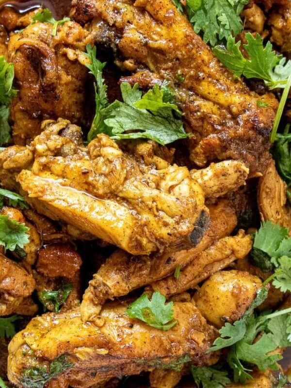 A plate of spiced, cooked chicken pieces garnished with fresh cilantro leaves, served with a spoon on a white, decorative dish.