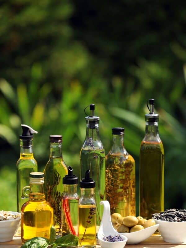 A selection of glass bottles filled with different oils, some infused with herbs and spices, displayed outdoors on a table with small bowls of olives, seeds, and fresh herbs in front, with greenery in the background.
