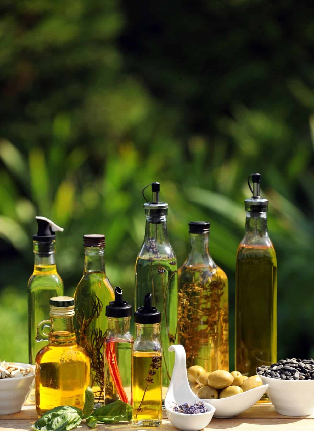 A selection of glass bottles filled with different oils, some infused with herbs and spices, displayed outdoors on a table with small bowls of olives, seeds, and fresh herbs in front, with greenery in the background.